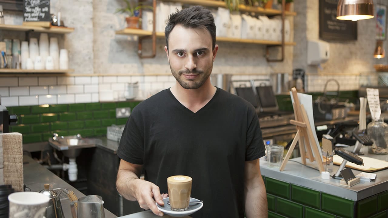 Alex in his coffee shop, holding a latte with pride, revealing how overcoming failure helped him build resilience for success